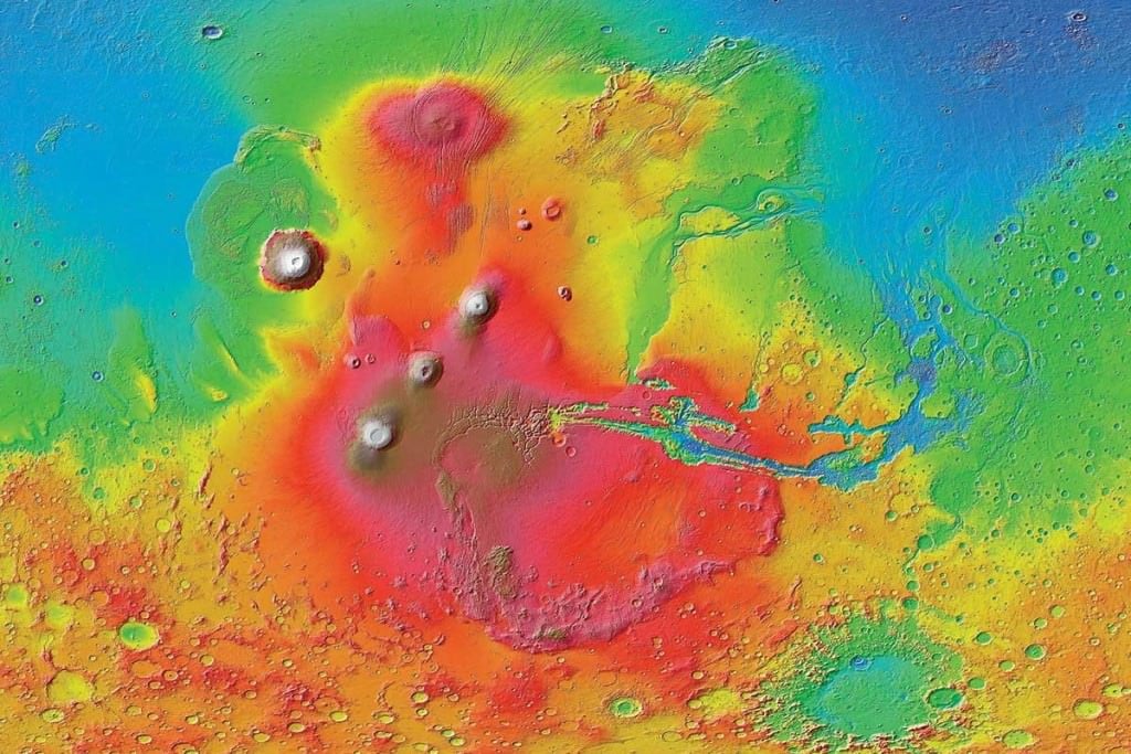A colorized image of the surface of Mars taken by the Mars Reconnaissance Orbiter. The line of three volcanoes is the Tharsis Montes, with Olympus Mons to the northwest. Valles Marineris is to the east. Image: NASA/JPL-Caltech/ Arizona State University